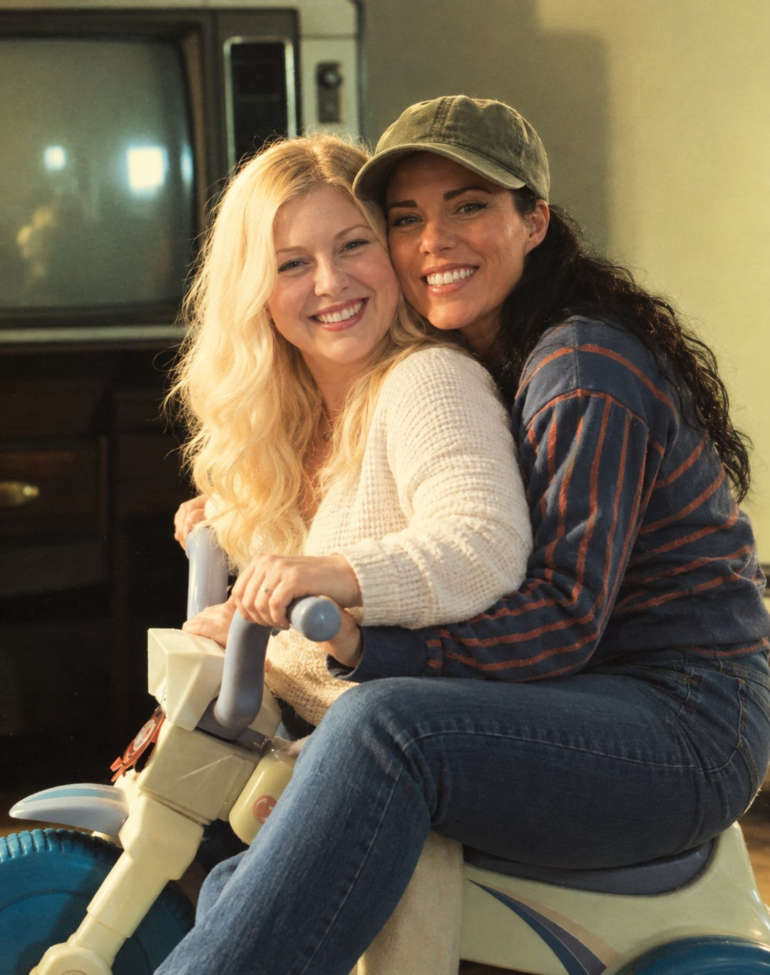 Kerri and Heather as adults, posed together on a childhood ride-on toy in a warmly lit living room